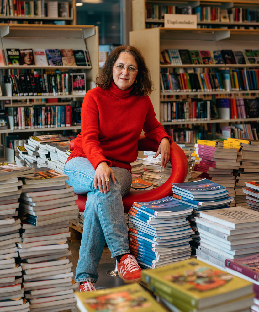 Ane Christiansen med rød genser på rød stol blant en haug med lærebøker