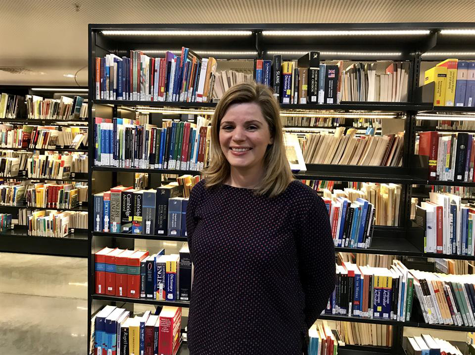 Edit Bugge står og smiler framfor ei bokhylle i eit bibliotek.