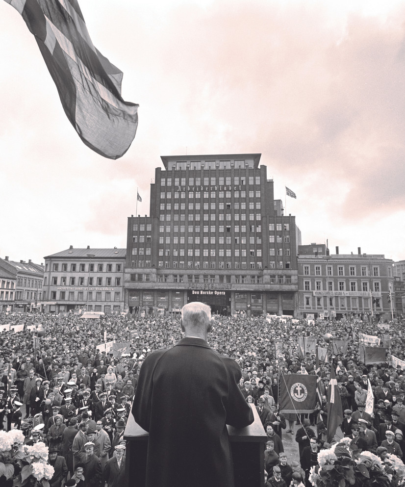 Einar Gerhardsen under vaiende norsk flagg på talerstolen på Youngstorget 1967, sett bakfra.