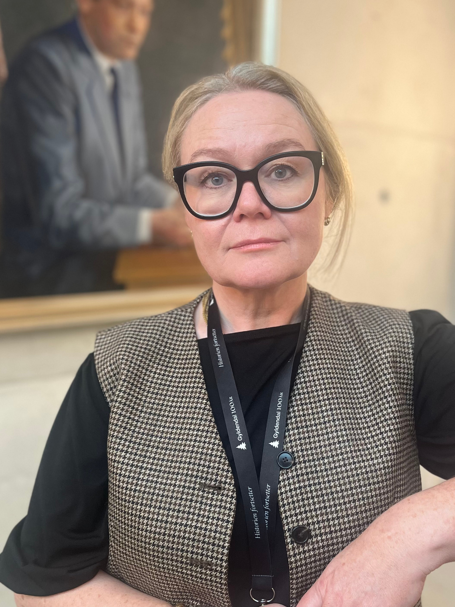 Hanne-Mari Eide Bennett har store briller med sort innfatning, adgangskort rundt halsen og et klassisk maleri i bakgrunnen.